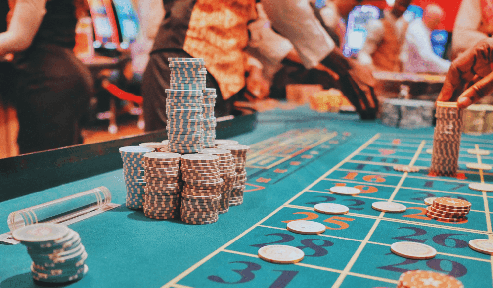 Poker chips on a green felted table, with casino dealers in the background. 