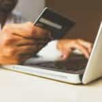 A dark-skinned man in a button down shirt holds a credit card next to his computer as he types.