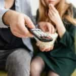 A man points a television remote, with popcorn and woman.