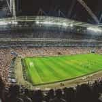 A fully packed-out soccer stadium during a soccer game, with the green pitch in the center, illuminated by bright floodlights.