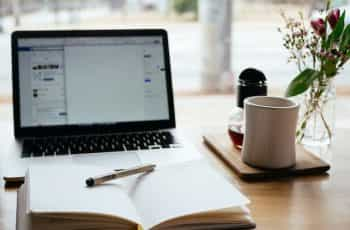 An open notebook lying flat on a table in front of a laptop next to a coffee mug and flower vase.