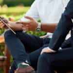 Two people in business wear sitting on a bench outside and discussing matters together, with one person showing information to the other on a tablet device.